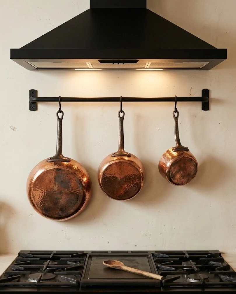 Three copper saucepans hung on a black rail above a kitchen range with a cream wall