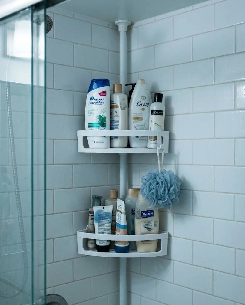 Corner tension-rod shower caddy wedged into a tiled shower corner holding shampoo bottles.