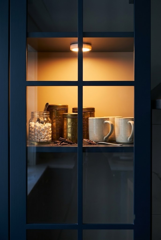 Glass-front cabinet shelf with cocoa tins and mugs lit by a warm battery puck light
